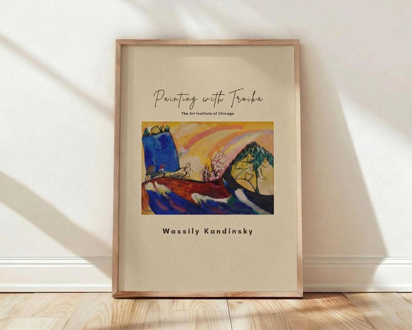 May include: Framed art print leaning against white wall on light wood floor with soft natural lighting and shadows. Features colorful abstract landscape with blue mountains, rainbow, tree, and geometric shapes in Kandinsky's style. Cream matting with wooden frame, styled in minimalist interior setting.