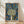 May include: Framed art print leaning against white wall on light wood flooring with palm leaf shadows. Features stylized tree with dotted circular canopy in green, blue and black dots on cream background. Deep blue starry night sky surrounds tree with golden yellow orbs and flowing white lines. Natural wood frame. Museum text at bottom in black letters.