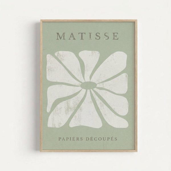 Framed poster with sage green background featuring a large white flower design in Matisse's cut-out style. The flower has eight rounded petals radiating from a central oval, with organic flowing shapes. Text reads "MATISSE" at top and "PAPIERS DÉCOUPÉS" at bottom in gray lettering. Natural wood frame against white wall background.