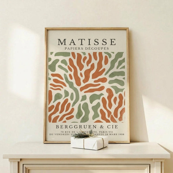 May include: Framed vintage-style art poster featuring abstract orange and sage green organic shapes in Matisse's cut-out style, displayed on light wood surface with small wrapped gift box, against cream wall in modern interior setting
