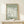 May include: Framed art print displayed on light wood surface showing large white flower silhouette on sage green background with "MATISSE" and "PAPIERS DÉCOUPÉS" text. Natural wood frame, small wrapped gift with twine beside it, white ceramic vase partially visible, neutral wall background with soft natural lighting.