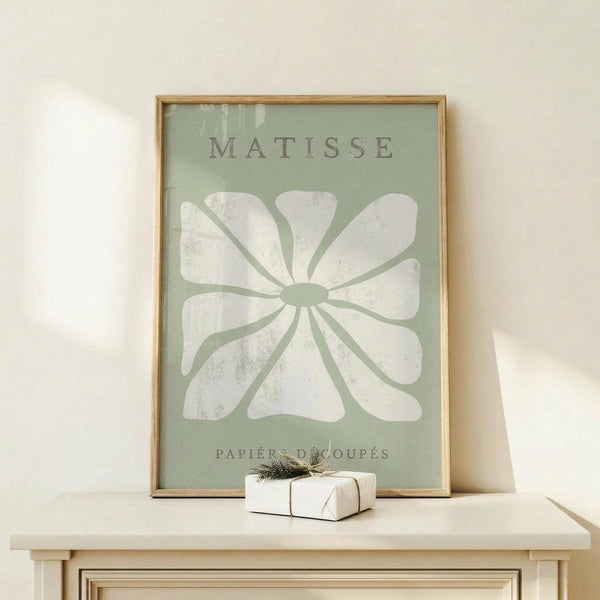 May include: Framed art print displayed on light wood surface showing large white flower silhouette on sage green background with "MATISSE" and "PAPIERS DÉCOUPÉS" text. Natural wood frame, small wrapped gift with twine beside it, white ceramic vase partially visible, neutral wall background with soft natural lighting.
