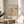 May include: Framed art print featuring abstract organic shapes in orange and green on cream background, displayed on round wooden stool beside beige upholstered chair. Natural sunlight creates soft shadows on white wall. Plant leaves visible in corner. Print shows cut-out paper style design with flowing, coral-like forms in warm autumn colors within thin wooden frame.