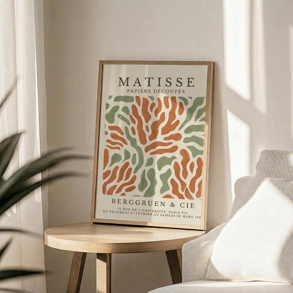 May include: Framed art print featuring abstract organic shapes in orange and green on cream background, displayed on round wooden stool beside beige upholstered chair. Natural sunlight creates soft shadows on white wall. Plant leaves visible in corner. Print shows cut-out paper style design with flowing, coral-like forms in warm autumn colors within thin wooden frame.