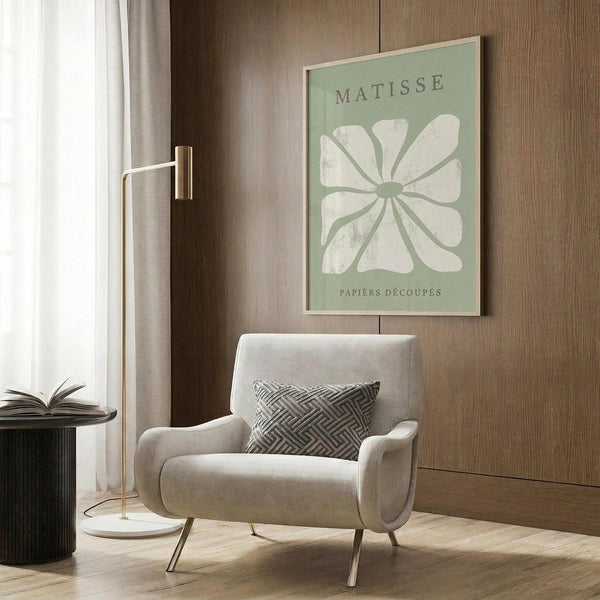 May include: Sage green and white Matisse-inspired botanical poster displayed in modern living room with beige upholstered armchair, geometric patterned pillow, brass floor lamp, and wood wall paneling. Abstract flower cutout design with white petals on muted green background in light wood frame.