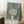 May include: Framed poster with stylized tree design featuring dotted circular canopy in green and blue tones, white trunk, swirling blue background with orange circles and white dots, text reading "Musee d'Art Moderne de la Ville de Paris" at bottom, displayed in light wood frame leaning against white wall next to wooden side table with coffee cup and cream textured throw blanket
