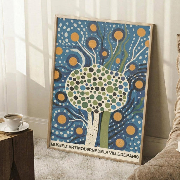 May include: Framed poster with stylized tree design featuring dotted circular canopy in green and blue tones, white trunk, swirling blue background with orange circles and white dots, text reading "Musee d'Art Moderne de la Ville de Paris" at bottom, displayed in light wood frame leaning against white wall next to wooden side table with coffee cup and cream textured throw blanket