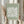 May include: Person in beige sweater holding framed art print with sage green background featuring white abstract flower or leaf pattern in Matisse cut-paper style. Natural wood frame, dried pampas grass visible on left side, soft neutral lighting near window with white curtains.