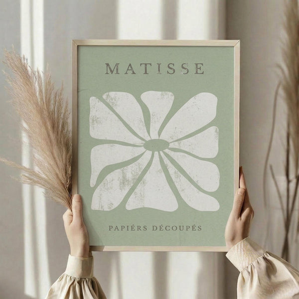 May include: Person in beige sweater holding framed art print with sage green background featuring white abstract flower or leaf pattern in Matisse cut-paper style. Natural wood frame, dried pampas grass visible on left side, soft neutral lighting near window with white curtains.