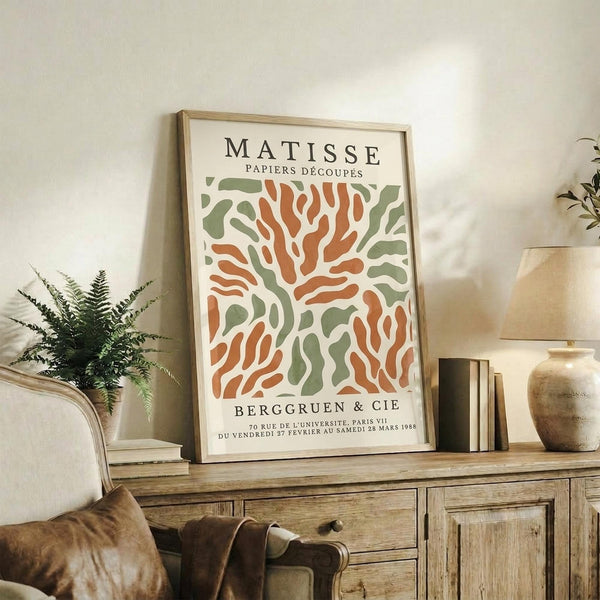 May include: Framed art print with abstract coral and seaweed-like shapes in orange and sage green on cream background, displayed on rustic wooden sideboard alongside potted fern, ceramic vase, stacked books, and table lamp in neutral-toned living room setting