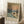 May include: Framed art print displaying an Impressionist winter river scene with sailing boats, bare trees, and warm sunset sky in muted blues, oranges and whites. The poster features "MONET" text at bottom and leans against white wall near wooden side table with coffee cup and cozy blanket in softly lit room.
