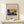 May include: Framed art print featuring a vibrant abstract painting with bold blues, oranges, and yellows depicting stylized boats or ships on water with geometric mountain forms in background. Light wood frame on cream background, displayed on pale wooden surface beside small white wrapped gift with twine and evergreen sprig. Clean minimalist setting with soft natural lighting.