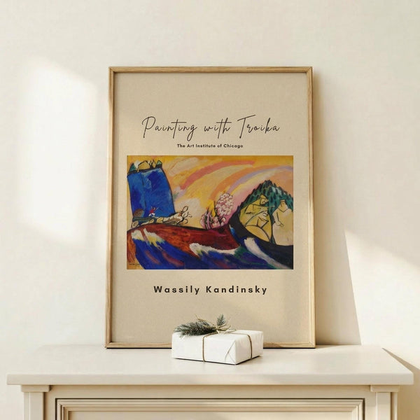 May include: Framed art print featuring a vibrant abstract painting with bold blues, oranges, and yellows depicting stylized boats or ships on water with geometric mountain forms in background. Light wood frame on cream background, displayed on pale wooden surface beside small white wrapped gift with twine and evergreen sprig. Clean minimalist setting with soft natural lighting.