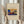 May include: Hands holding a framed art print featuring an abstract landscape with vibrant blue, yellow, orange and red colors, mountains, rainbow, and pink flowering tree. Light wood frame against cream mat. Person wearing beige ruffled sleeves holds dried pampas grass. Soft neutral interior with white curtains in background.