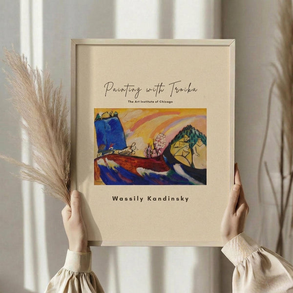 May include: Hands holding a framed art print featuring an abstract landscape with vibrant blue, yellow, orange and red colors, mountains, rainbow, and pink flowering tree. Light wood frame against cream mat. Person wearing beige ruffled sleeves holds dried pampas grass. Soft neutral interior with white curtains in background.