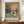 May include: Framed Monet-style winter landscape poster showing snow-covered riverbank with bare trees, sailboat on water during sunset or sunrise with peachy-orange sky. Print displayed on wooden sideboard next to white ceramic vase and stacked books in elegant room with cream walls and white molding.