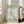 May include: Large framed poster with sage green background featuring white abstract flower cutout design in Matisse style, displayed against white wall corner next to beige upholstered chair in bright sunlit room with wooden floors and white window frames
