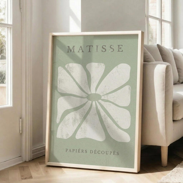 May include: Large framed poster with sage green background featuring white abstract flower cutout design in Matisse style, displayed against white wall corner next to beige upholstered chair in bright sunlit room with wooden floors and white window frames