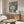May include: Modern art poster featuring a stylized tree with circular dotted foliage in green tones against a blue background with white dots and golden yellow circles, displayed above a mid-century walnut credenza in a contemporary living room with white sculptural chair and glass coffee table.
