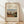 May include: Framed art print featuring Henri Rousseau's painting of a Parisian suburban street scene with small houses, red tile roofs, trees, and small figures walking on an earthen road under cloudy skies. The poster has cream matting with black text reading "OUTSKIRTS OF PARIS" and artist attribution. Natural wood frame sits against white wall on light wooden floor with soft natural lighting creating diagonal shadows.