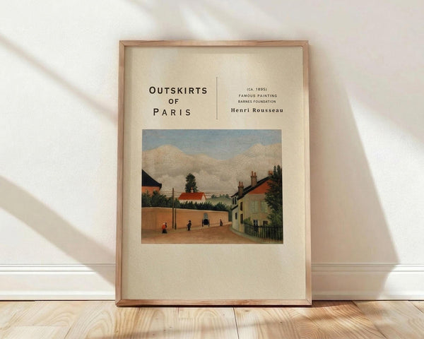 May include: Framed art print featuring Henri Rousseau's painting of a Parisian suburban street scene with small houses, red tile roofs, trees, and small figures walking on an earthen road under cloudy skies. The poster has cream matting with black text reading "OUTSKIRTS OF PARIS" and artist attribution. Natural wood frame sits against white wall on light wooden floor with soft natural lighting creating diagonal shadows.