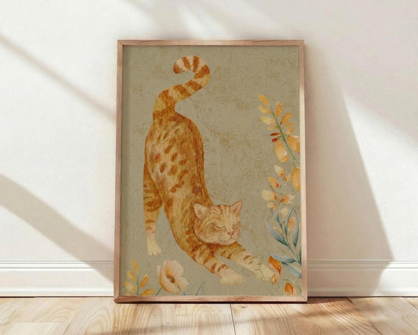 May include: Framed watercolor artwork showing an orange tabby cat in a stretching pose with its back arched and tail curved upward. The cat has distinctive striped markings in warm orange and cream tones. Delicate botanical elements with peachy flowers and green stems accent the composition against a soft sage green background. The piece is displayed in a thin wooden frame, leaning against a white wall on light wooden flooring with natural sunlight creating soft shadows.