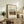 May include: Framed art print displaying Henri Rousseau's pastoral village scene with houses, mountains, and figures on a dirt path. The poster features cream matting with black text reading "Outskirts of Paris" and artist details. Styled on rustic wooden sideboard alongside fern plant, ceramic vase, stacked books, and cream upholstered chair with brown leather pillow in neutral-toned living room.
