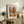 May include: Framed Matisse-style art poster displayed on rustic wooden sideboard in cozy living room setting. Colorful still life artwork features vibrant flowers in geometric vase with oranges, set against blue and coral background. Natural wood frame complements warm interior with fern plant, ceramic pottery, books, and cream upholstered furniture visible.