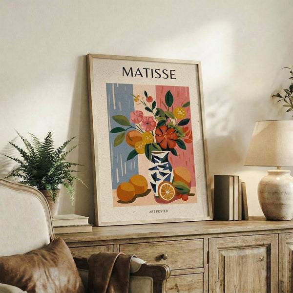 May include: Framed Matisse-style art poster displayed on rustic wooden sideboard in cozy living room setting. Colorful still life artwork features vibrant flowers in geometric vase with oranges, set against blue and coral background. Natural wood frame complements warm interior with fern plant, ceramic pottery, books, and cream upholstered furniture visible.