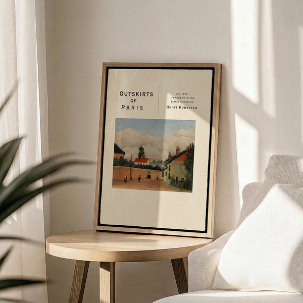 May include: Framed art print featuring Henri Rousseau's pastoral village scene with houses, trees, and small figures under a cloudy sky, displayed on a light wood round side table next to a beige upholstered chair with white throw pillow. Soft natural lighting creates shadows on the neutral wall behind. The poster shows "Outskirts of Paris" text above the colorful landscape painting in an exhibition-style layout with cream background and thin wood frame.