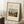 May include: Framed art print leaning against white wall showing a village scene with houses, trees, and small figures under cloudy sky. Cream-colored poster with black text reading "OUTSKIRTS OF PARIS" and "Henri Rousseau" above the artwork. Natural wood frame with black border. Coffee cup on wooden block nearby, beige textured rug below, soft natural lighting creating shadows.