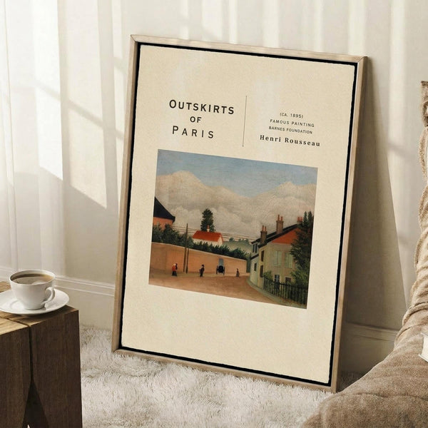 May include: Framed art print leaning against white wall showing a village scene with houses, trees, and small figures under cloudy sky. Cream-colored poster with black text reading "OUTSKIRTS OF PARIS" and "Henri Rousseau" above the artwork. Natural wood frame with black border. Coffee cup on wooden block nearby, beige textured rug below, soft natural lighting creating shadows.
