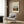 May include: Framed art print featuring a pastoral village scene with buildings, trees, and cloudy sky in muted earth tones. Print hangs on wood-paneled wall above a cream upholstered armchair with geometric patterned pillow. Modern brass floor lamp and dark round side table with open book complete the contemporary reading nook scene.