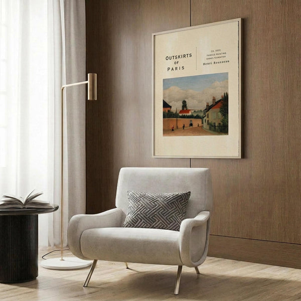 May include: Framed art print featuring a pastoral village scene with buildings, trees, and cloudy sky in muted earth tones. Print hangs on wood-paneled wall above a cream upholstered armchair with geometric patterned pillow. Modern brass floor lamp and dark round side table with open book complete the contemporary reading nook scene.
