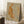 May include: Framed watercolor artwork featuring an orange tabby cat stretching with curved back and raised tail, surrounded by delicate botanical elements in warm autumn tones. The piece has a soft, muted background and is displayed in a light wooden frame, leaning against a white wall beside a cozy reading nook with coffee cup, books, and textured throw blanket on plush carpet.