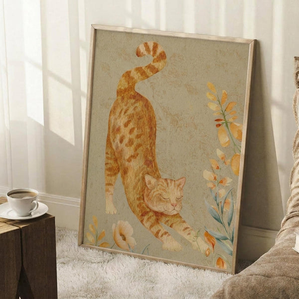 May include: Framed watercolor artwork featuring an orange tabby cat stretching with curved back and raised tail, surrounded by delicate botanical elements in warm autumn tones. The piece has a soft, muted background and is displayed in a light wooden frame, leaning against a white wall beside a cozy reading nook with coffee cup, books, and textured throw blanket on plush carpet.