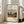 May include: Framed art print leaning against white wall next to beige upholstered chair in bright room with large windows. Print shows pastoral village scene with orange-roofed houses, small figures walking on dirt path, green trees and cloudy sky. Text reads "Outskirts of Paris" and "Henri Rousseau" with museum-style formatting on cream background. Light wood frame with black mat border.