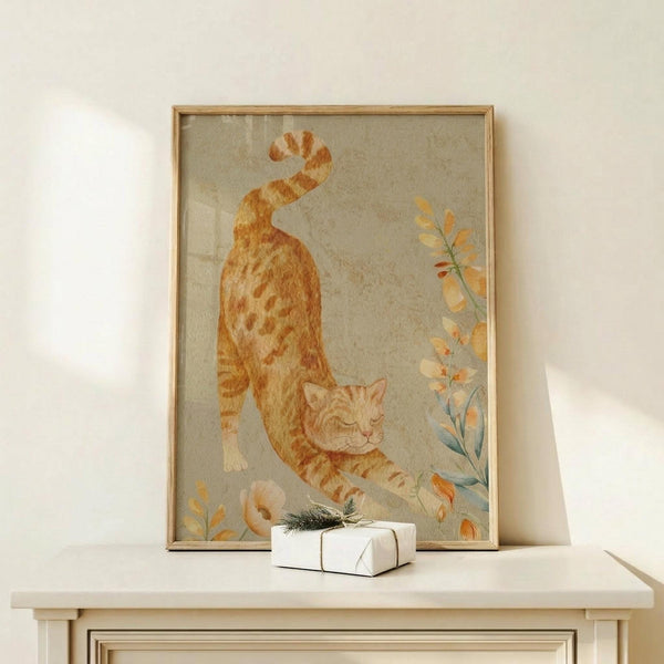 May include: Framed watercolor artwork showing an orange tabby cat stretching in a downward pose, surrounded by delicate botanical elements with orange and yellow leaves on a soft sage green background. The piece is displayed on a light wood console table beside a small wrapped gift with twine and evergreen sprig. The frame appears natural wood toned against a cream colored wall with soft natural lighting.