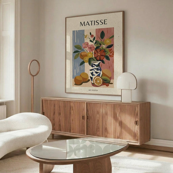 May include: Framed art poster displayed above wooden sideboard in modern living room with white curved chair and glass coffee table. Poster features colorful still life with flowers in striped vase and oranges in Matisse's distinctive cut-out style, with blue, pink, and coral background sections. Room has neutral walls, natural lighting, and contemporary furniture with warm wood tones.