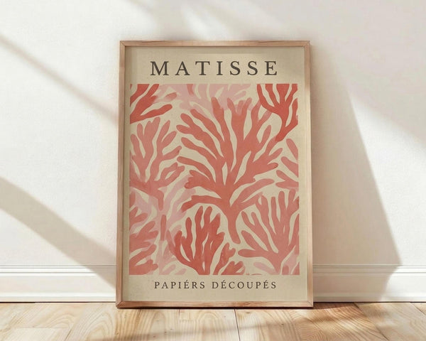 May include: Framed art print featuring coral-colored abstract cut-out shapes resembling flowing seaweed or coral branches against cream background, with "MATISSE" and "PAPIERS DÉCOUPÉS" text. Natural wood frame leaning against white wall on light wooden floor with soft window light creating diagonal shadows across white fabric backdrop.