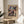 May include: Framed Paul Klee exhibition poster with abstract geometric artwork in muted colors including blues, purples, pinks and earth tones, displayed on a round wooden side table next to a beige upholstered chair with a throw pillow, palm plant visible in corner, soft natural lighting creating shadows on white walls