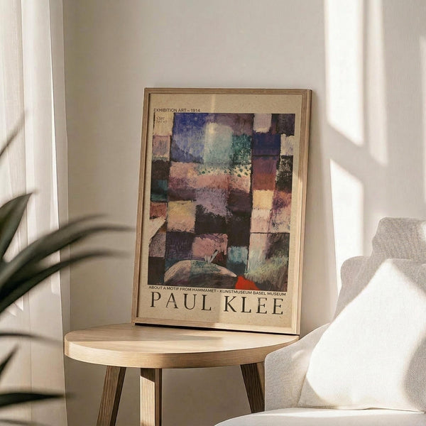 May include: Framed Paul Klee exhibition poster with abstract geometric artwork in muted colors including blues, purples, pinks and earth tones, displayed on a round wooden side table next to a beige upholstered chair with a throw pillow, palm plant visible in corner, soft natural lighting creating shadows on white walls