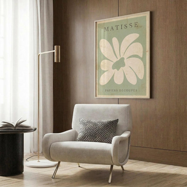 May include: Framed Matisse-inspired poster with sage green background and cream colored abstract flower cutout design, displayed on wood paneled wall above beige upholstered armchair with geometric patterned pillow, brass floor lamp and side table with open book nearby
