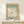 May include: Framed vintage-style poster with sage green background featuring large white flower cutout design in Matisse's paper collage style, displayed on light wood surface with small wrapped gift box, leaning against cream wall in minimalist setting