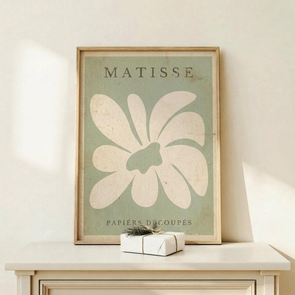 May include: Framed vintage-style poster with sage green background featuring large white flower cutout design in Matisse's paper collage style, displayed on light wood surface with small wrapped gift box, leaning against cream wall in minimalist setting