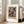 May include: A framed Paul Klee exhibition poster featuring an abstract geometric composition with colorful squares and rectangles in blues, greens, purples, and earth tones on a beige background. The artwork is displayed in a light wood frame with white matting, leaning against a white wall next to a beige upholstered chair in a bright, minimalist living room with large windows and natural light.