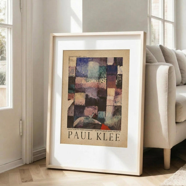May include: A framed Paul Klee exhibition poster featuring an abstract geometric composition with colorful squares and rectangles in blues, greens, purples, and earth tones on a beige background. The artwork is displayed in a light wood frame with white matting, leaning against a white wall next to a beige upholstered chair in a bright, minimalist living room with large windows and natural light.