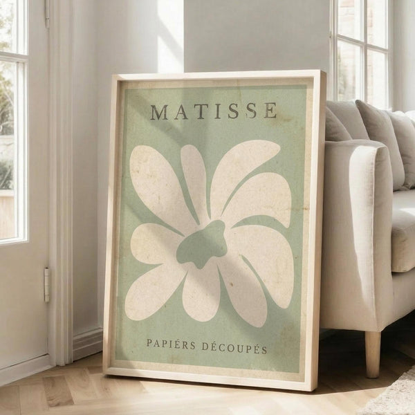 May include: Framed poster with sage green background featuring large white abstract flower shape in Matisse cut-out style, displayed leaning against white wall next to beige upholstered chair in bright living room with large windows and natural lighting