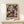 May include: Framed Paul Klee exhibition poster leaning against cream wall on light wood console table. Abstract artwork features geometric patches in muted blues, purples, pinks, and earth tones with mosaic-like composition. Small white wrapped gift with green sprig sits beside the thin gold frame. Soft natural lighting creates gentle shadows on minimalist surface.