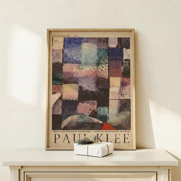 May include: Framed Paul Klee exhibition poster leaning against cream wall on light wood console table. Abstract artwork features geometric patches in muted blues, purples, pinks, and earth tones with mosaic-like composition. Small white wrapped gift with green sprig sits beside the thin gold frame. Soft natural lighting creates gentle shadows on minimalist surface.